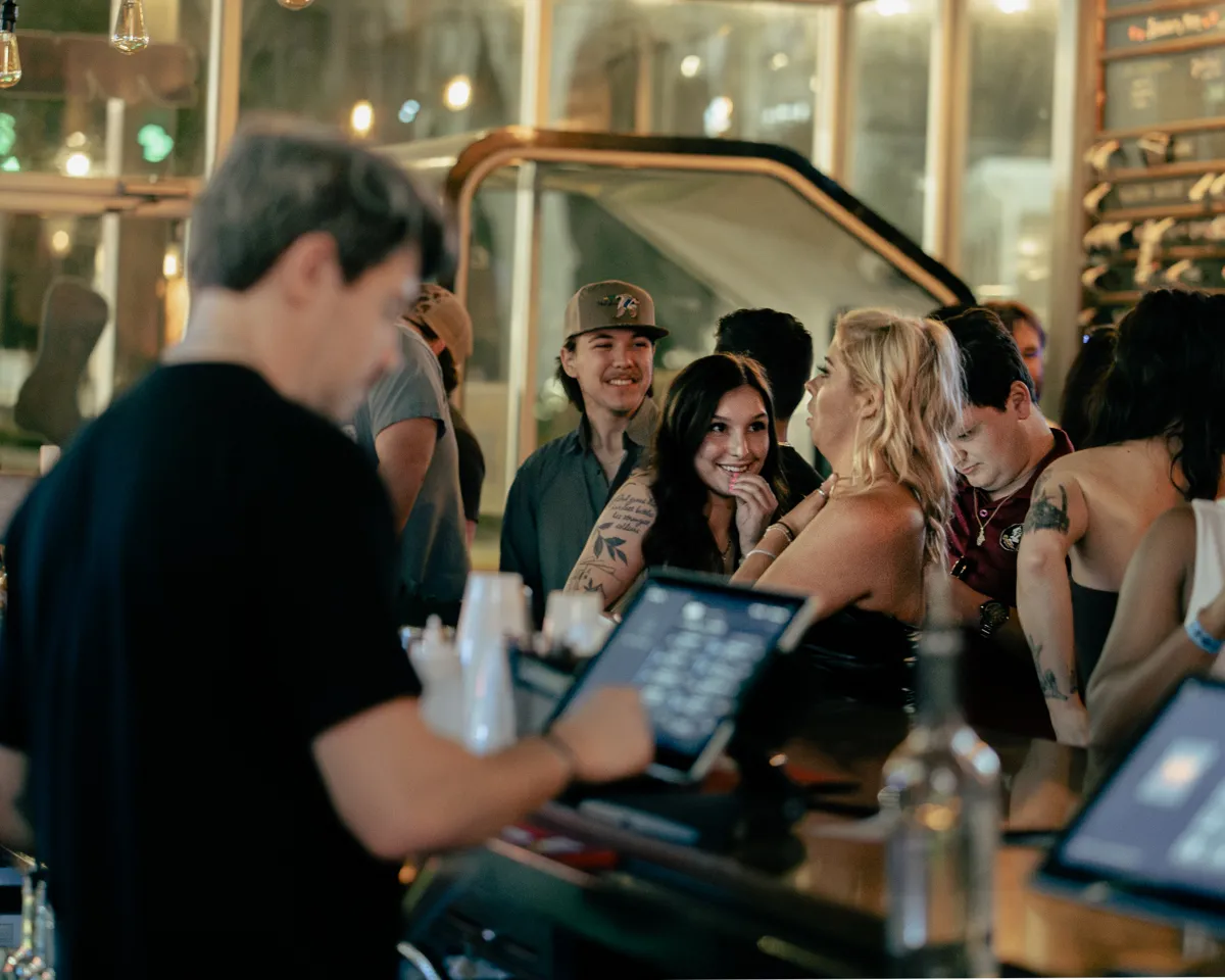 A bartender ringing in an order on a POS tablet while guests wait at a busy bar