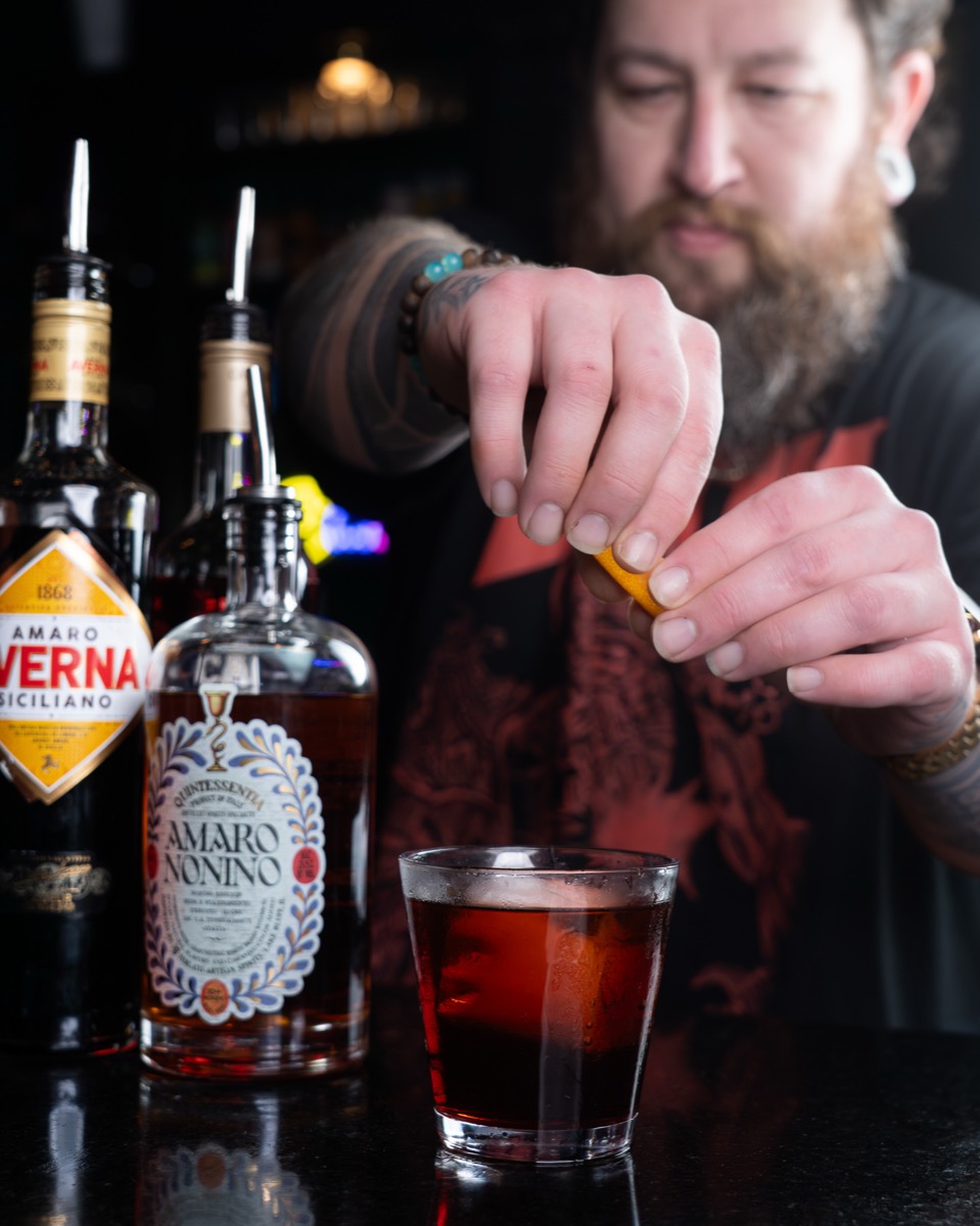 A bartender crafting a cocktail with amaro bottles on the bar