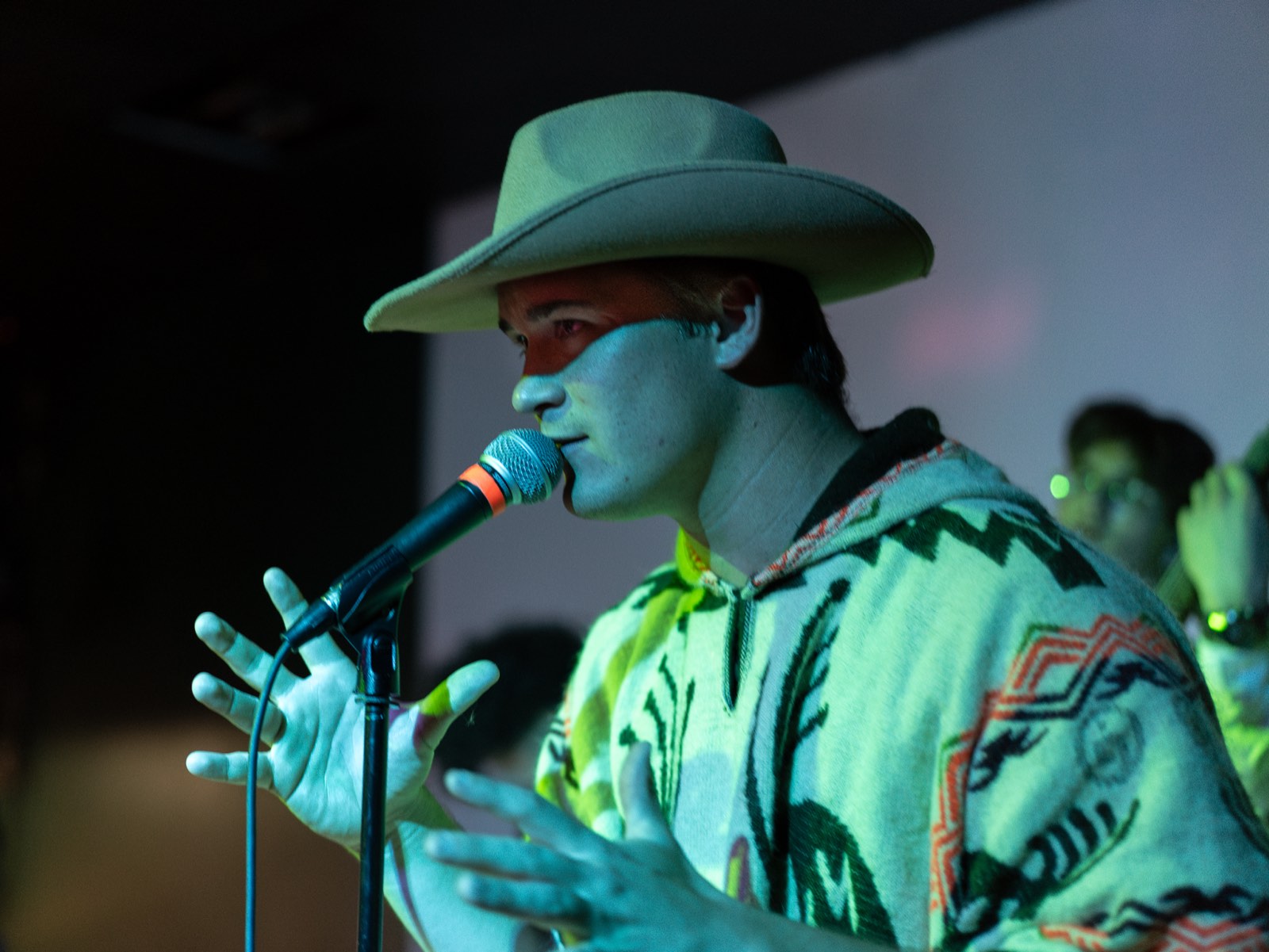 A performer in a cowboy hat singing into a mic under green stage lighting at a Denver bar